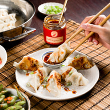 Load image into Gallery viewer, Person using chopsticks to pick up a dumpling from a plate with other food items on a wooden table.